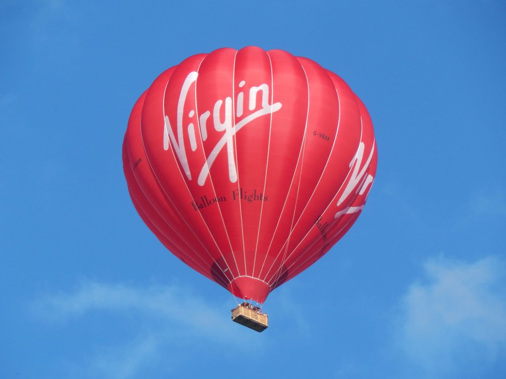 A red Virgin branded hot air balloon in the air, against a light blue sky.
